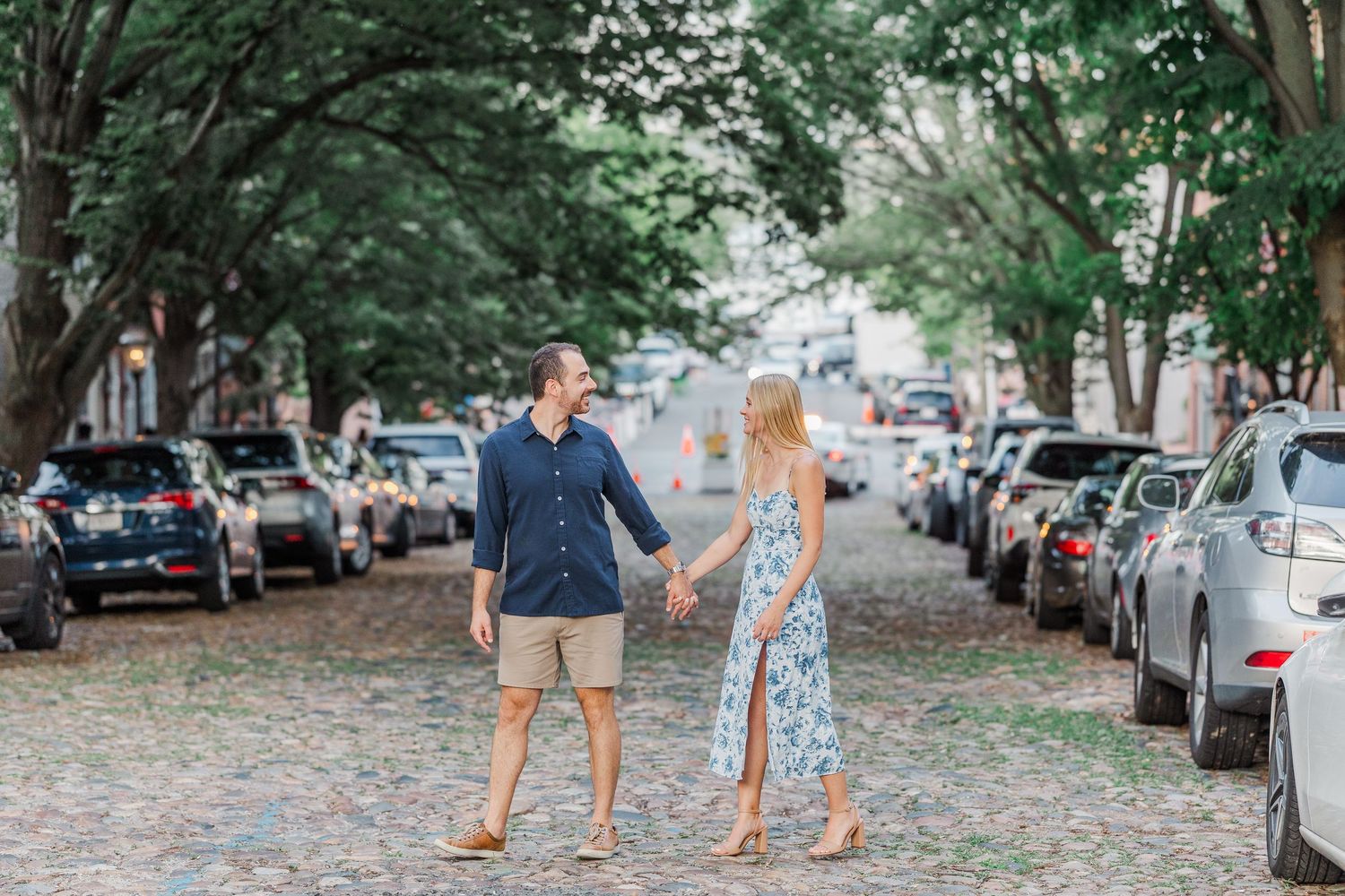 Couple holding hands on a charming cobblestone street lined with trees and parked cars in a romantic urban setting.