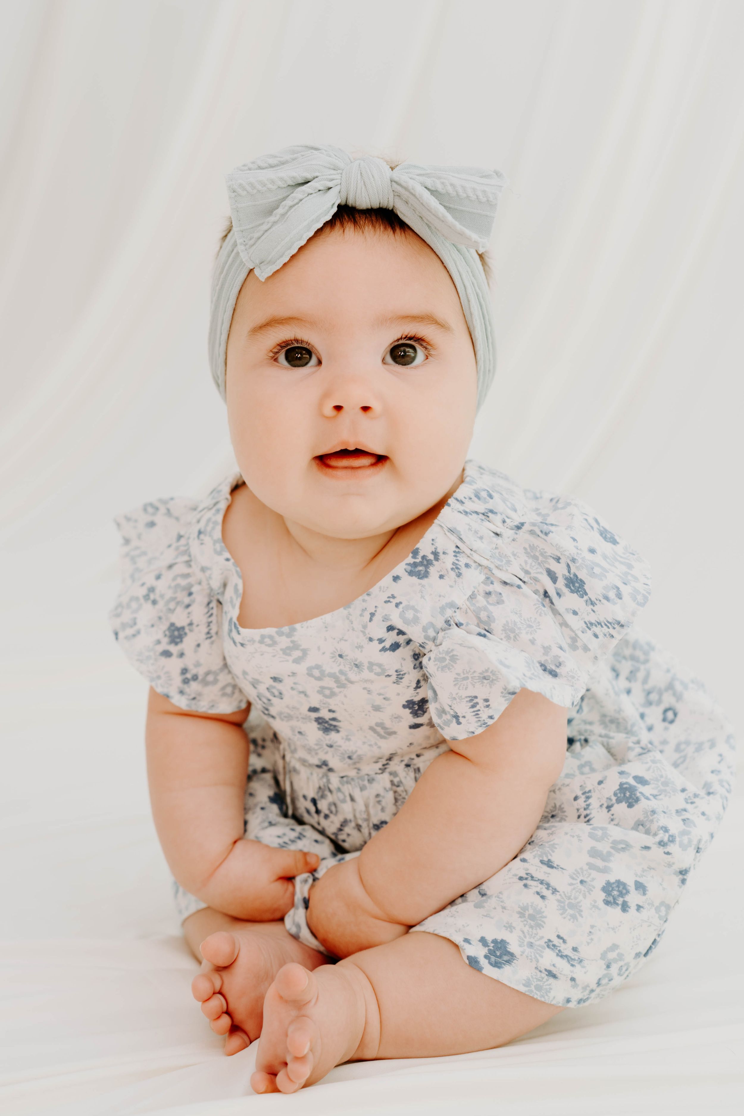 Adorable baby girl in floral romper and gray bow headband sitting up and looking at camera.