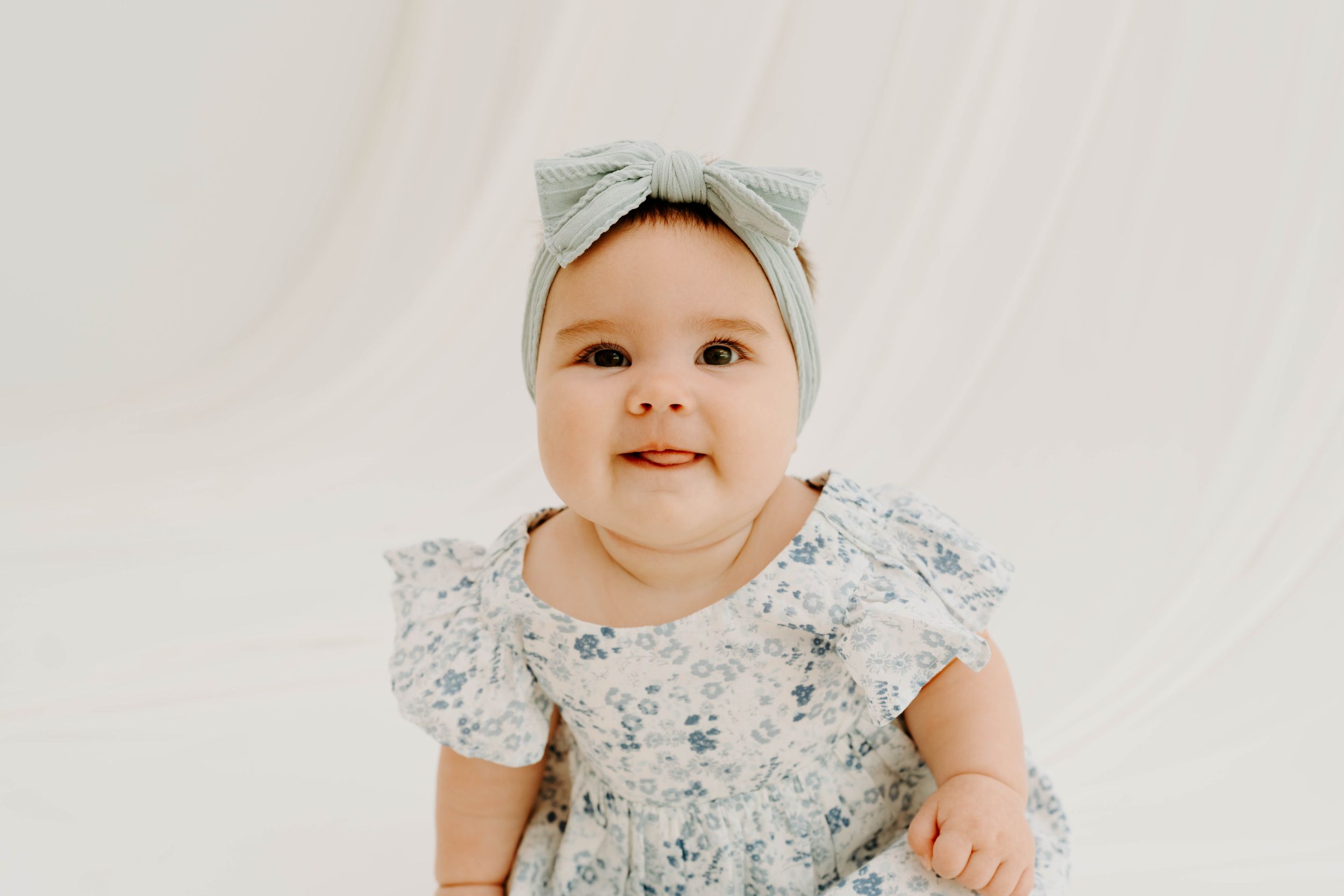 Smiling baby girl in floral dress and sage green bow headband poses for a portrait photo.