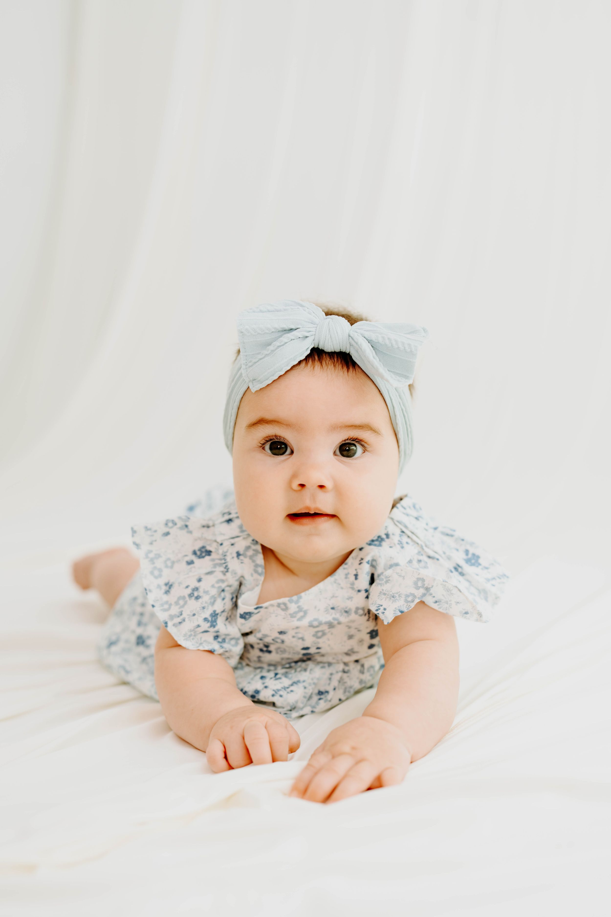 Adorable baby girl in a floral romper and blue bow headband doing tummy time on a white backdrop.