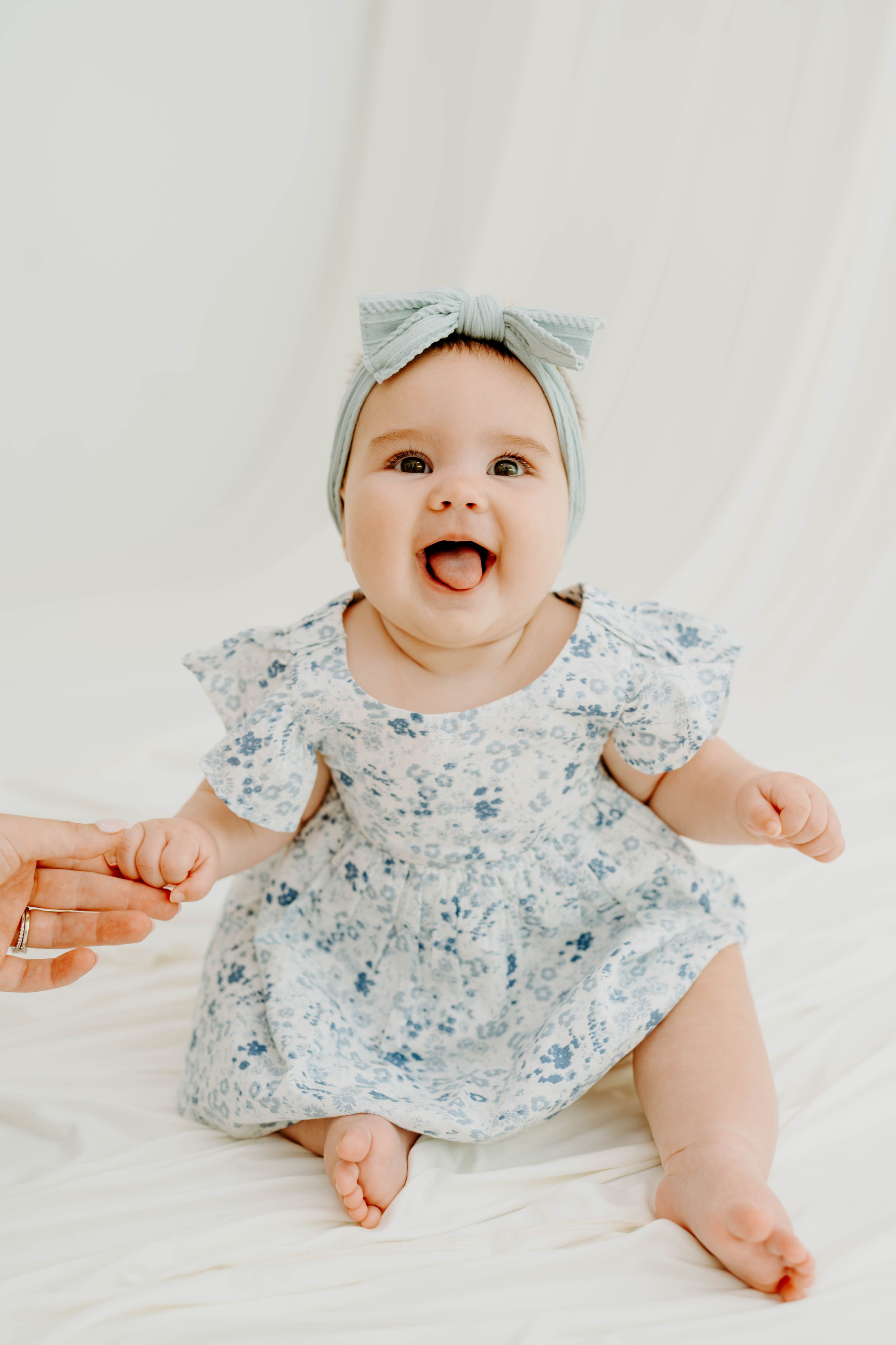 Adorable baby girl in floral dress and blue bow headband sitting and smiling with mouth open on white background.