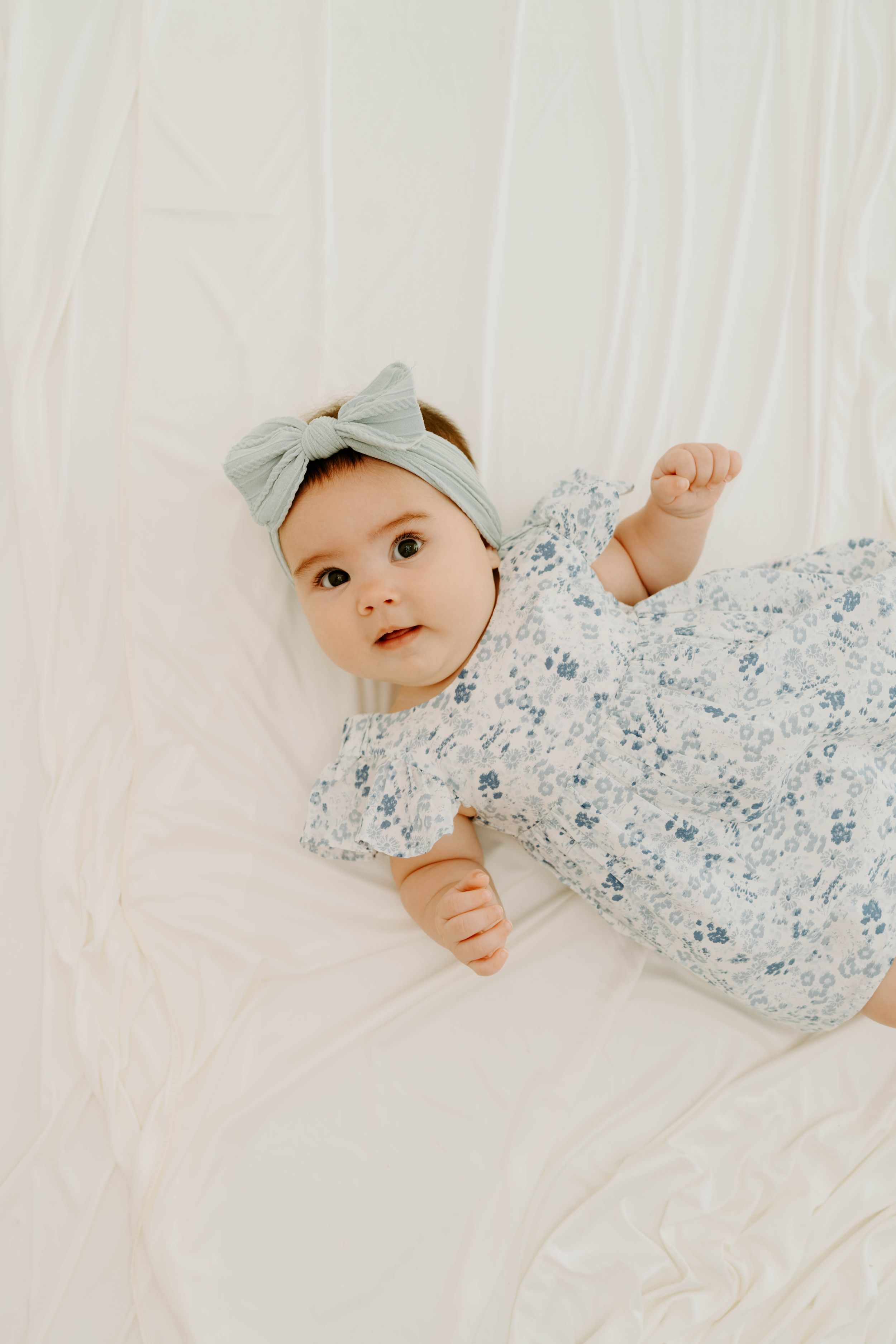 Adorable baby girl in floral romper and sage bow headband lying on white bedding, looking up curiously.