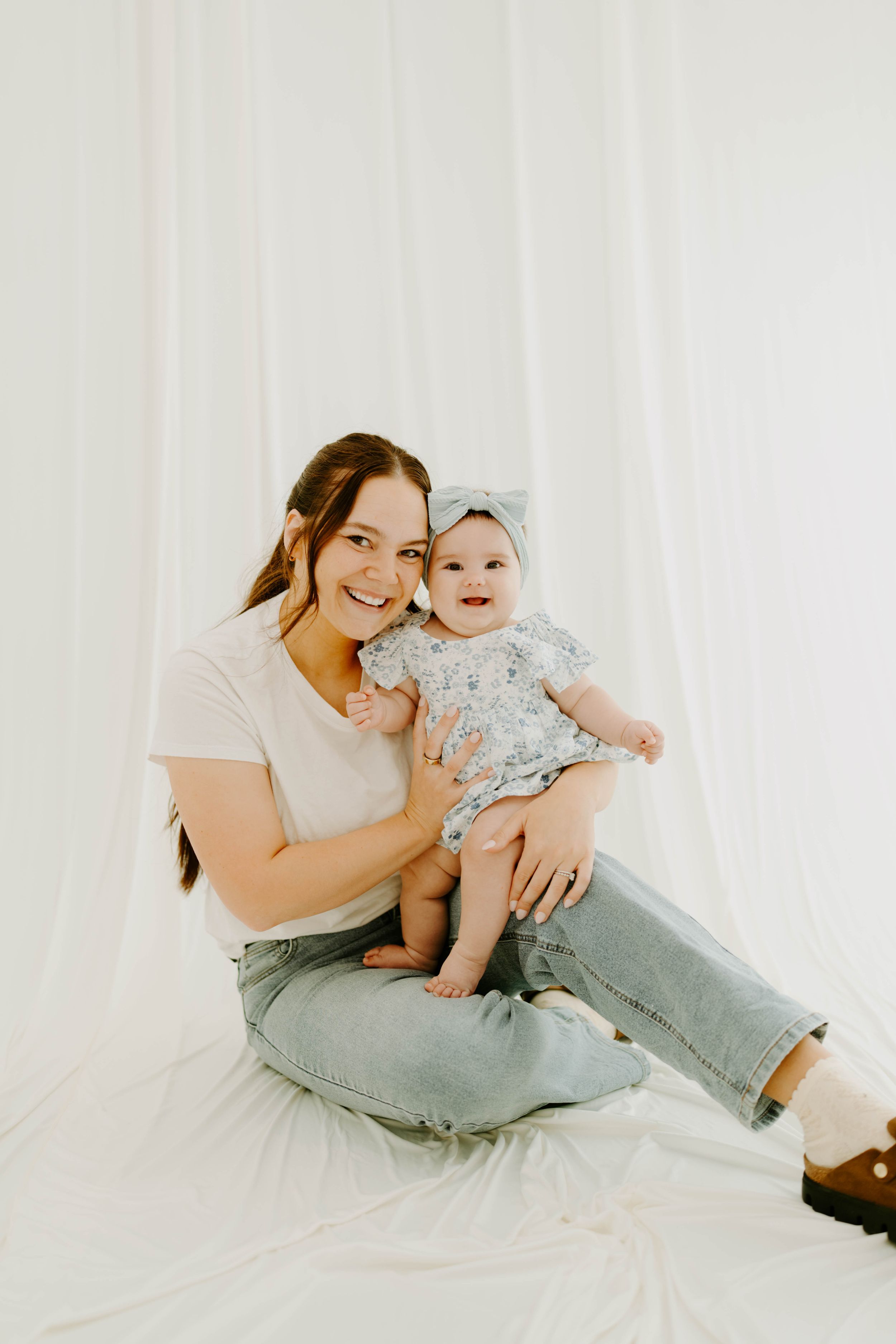 A young mother sits smiling on white fabric holding her happy baby girl in a floral outfit with a bow headband.