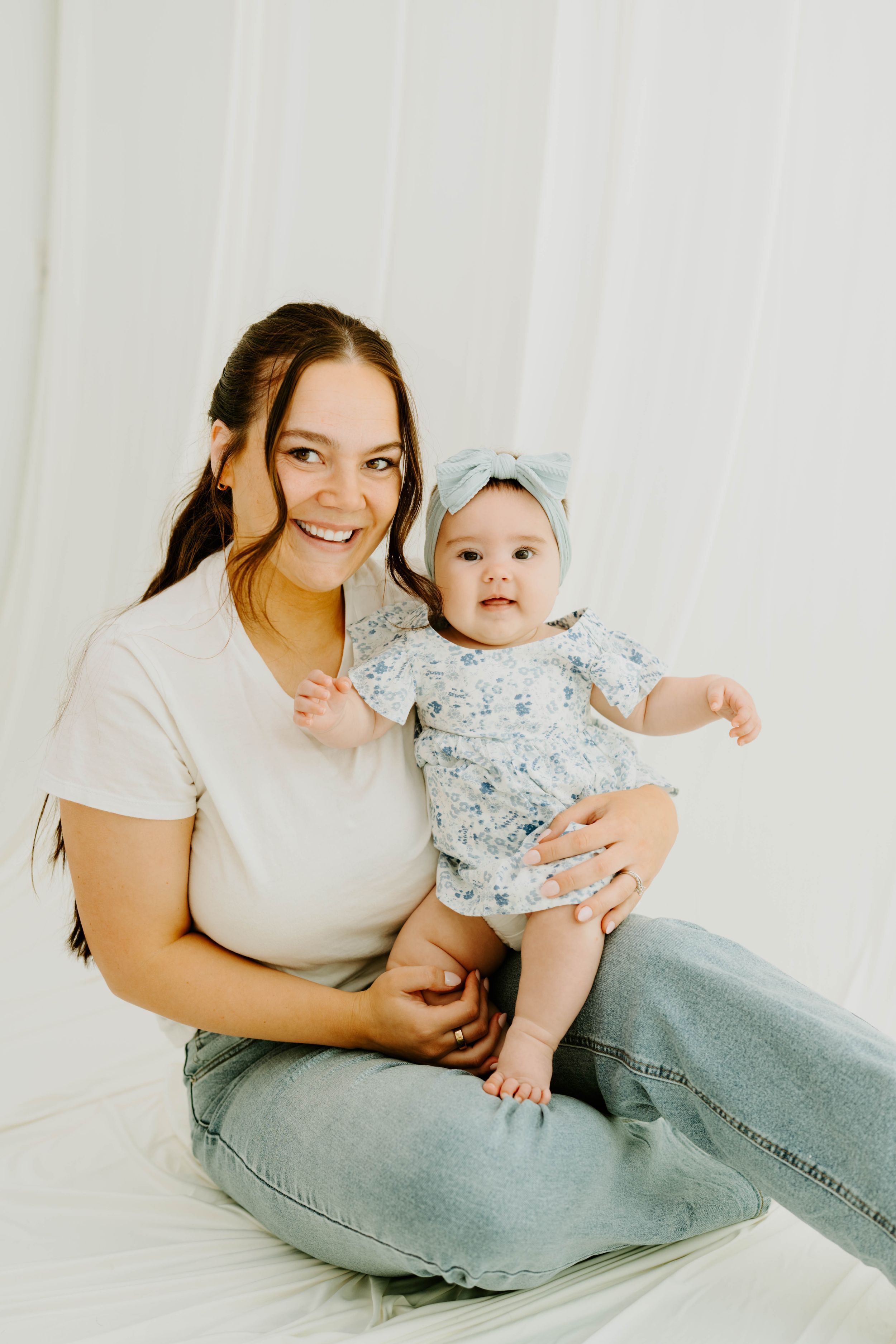 Smiling young mother sitting with her baby girl in a floral outfit and bow headband against a white backdrop.