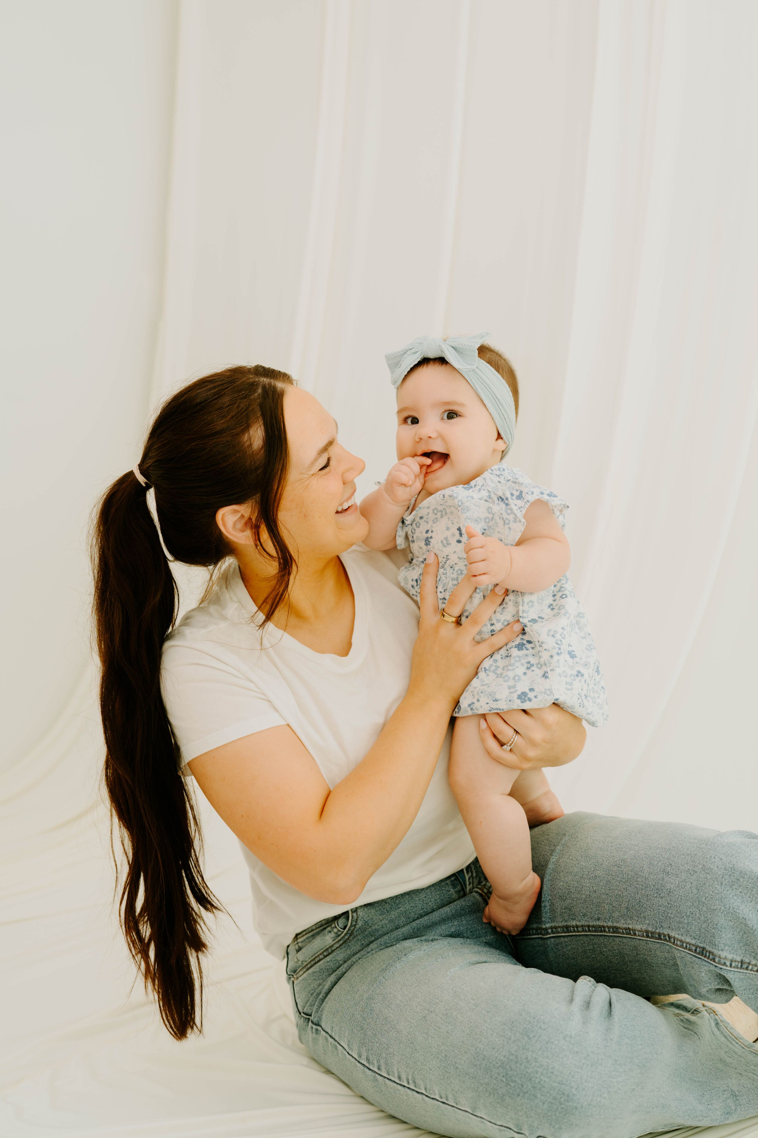 A smiling mother in a white tee holds her laughing baby girl in a floral outfit and bow headband against a white backdrop.