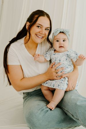 Smiling woman in white tee and jeans holds adorable baby girl in floral dress with blue bow headband.