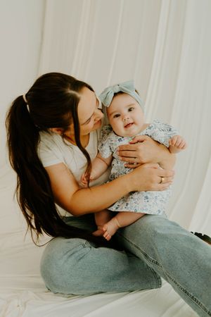 A mother with dark hair sits holding her smiling baby girl in a floral outfit and blue bow headband.