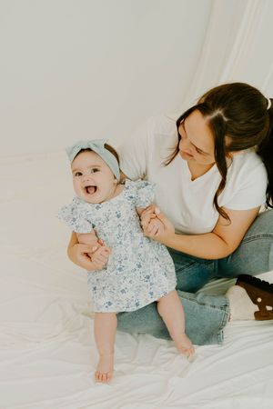 A smiling baby in a floral dress and bow headband stands with help from her dark-haired mother on a white backdrop.