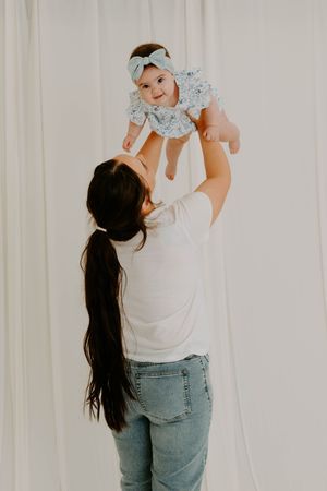 A mother lifts her smiling baby girl wearing a floral outfit and bow headband against a white backdrop.