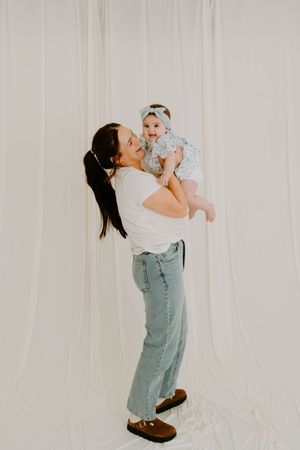A brunette mother lifts her baby girl overhead, both smiling, against a white curtain backdrop.