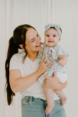 Smiling mother in white tee and jeans holds laughing baby girl in floral dress and blue bow headband.