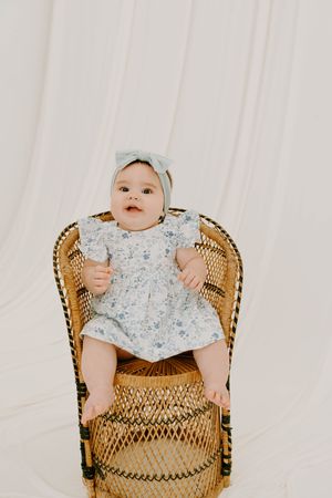 Baby girl in floral dress and bow headband sitting in wicker chair against white backdrop.