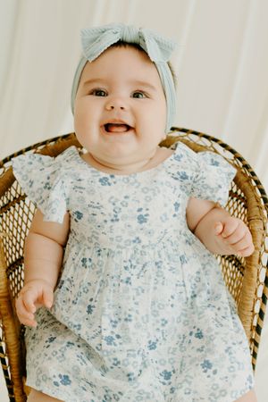 Smiling baby girl in white floral dress and blue bow headband sitting in wicker chair.