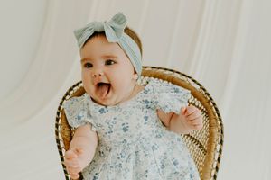 Happy baby girl in floral dress and mint bow headband sitting in wicker chair, mouth open with joy.