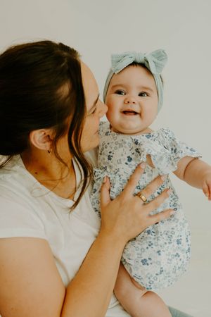 A mother kisses her happy baby girl in a floral romper and mint bow headband while holding her close.