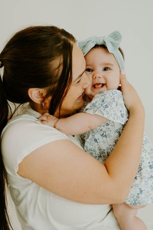 Loving mother kissing smiling baby girl in floral romper with blue bow headband against white background.