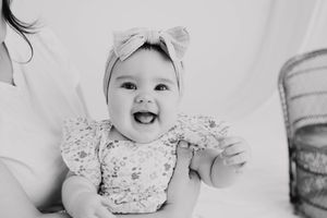 Smiling baby girl in floral romper and bow headband laughing joyfully in black and white portrait photo.