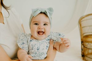 Smiling baby girl wearing a floral romper and sage green bow headband, held by mother in a bright, airy studio setting.