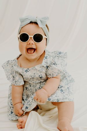 Adorable baby girl in floral dress and blue bow headband wearing round sunglasses, laughing joyfully.