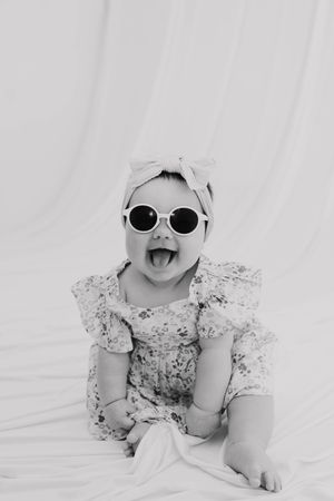 Black and white photo of a cute baby in floral romper and bow headband wearing round sunglasses, smiling with mouth open.