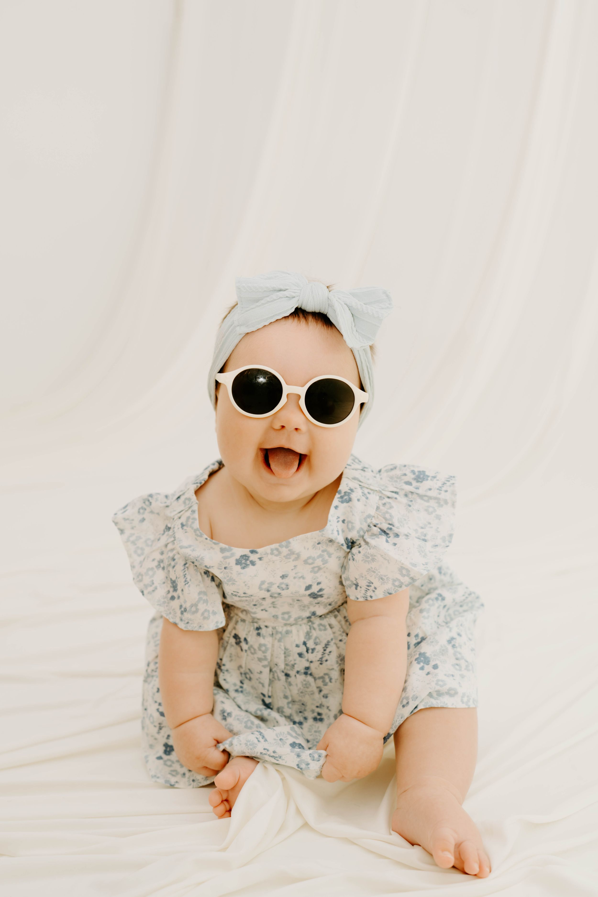 Happy baby girl in floral romper and blue headband wearing white round sunglasses, sitting and smiling.