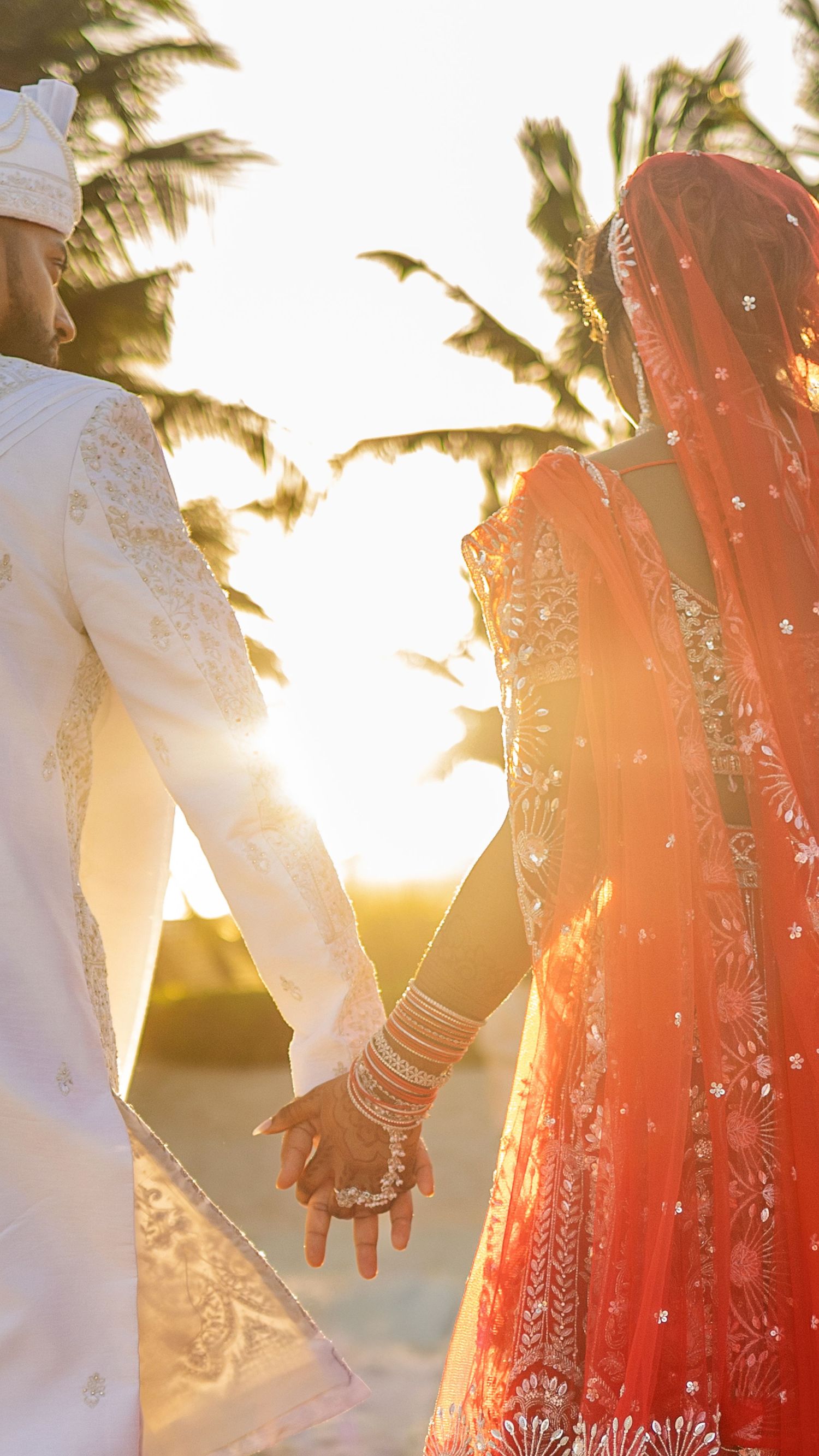 Indian bride in red lehenga and groom in white sherwani hold hands at sunset during traditional wedding ceremony.