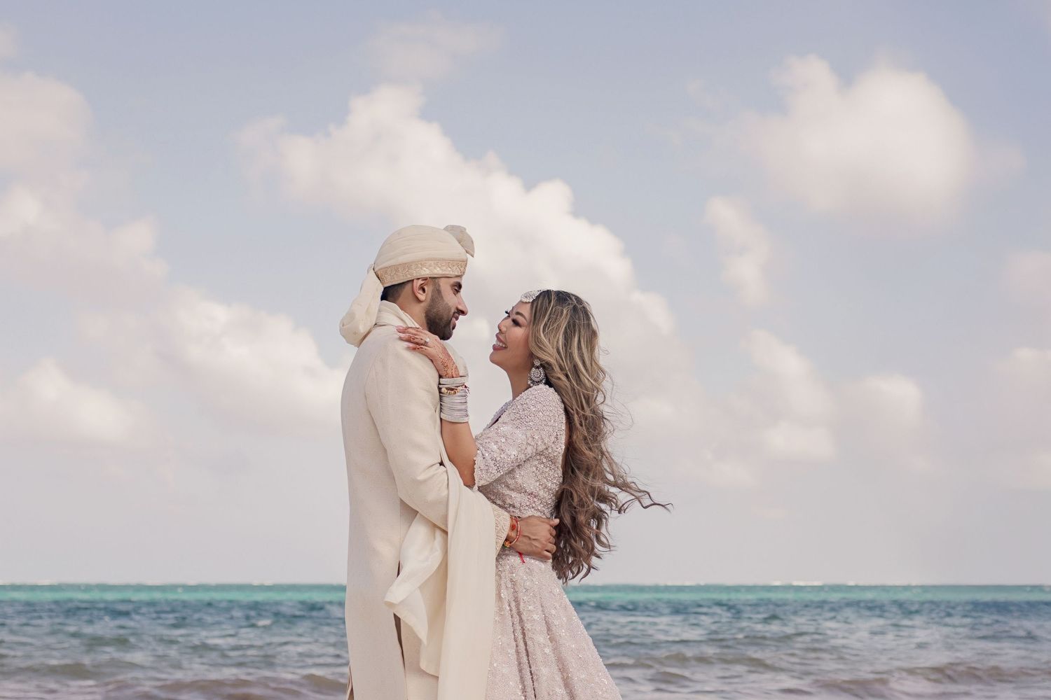 Indian couple in traditional wedding attire embracing on a tropical beach with turquoise ocean backdrop.