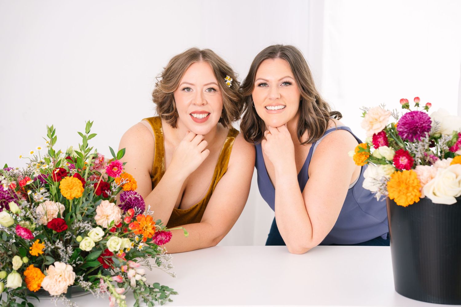 Two smiling women posing together behind colorful flower arrangements on white table.