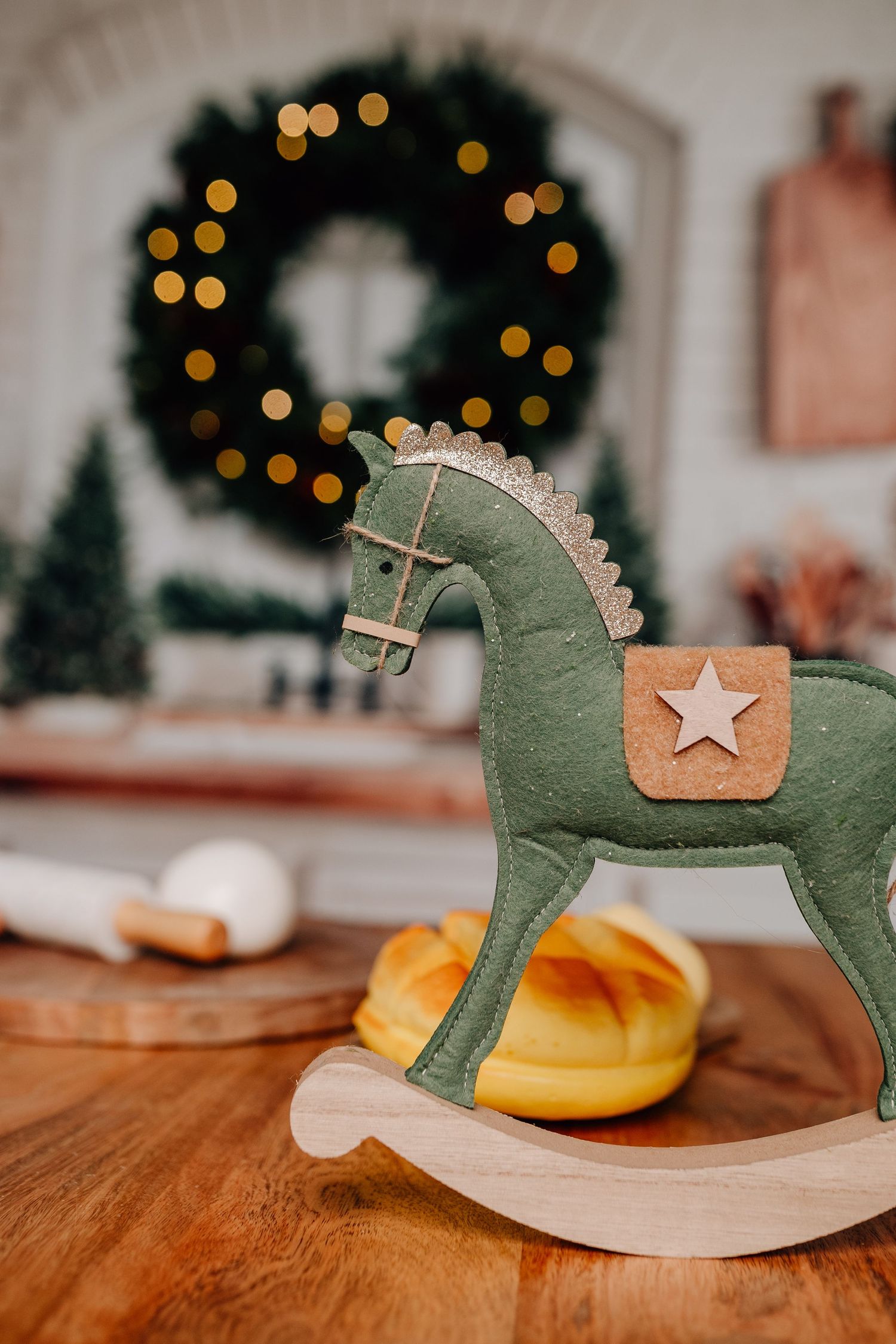 Christmas kitchen scene with rocking horse, bread, and vegetables on wooden board with festive tree decorations in background.