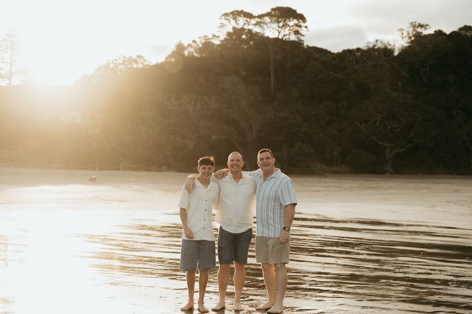Three men smiling together on a sunlit beach at golden hour with lush trees in the background.