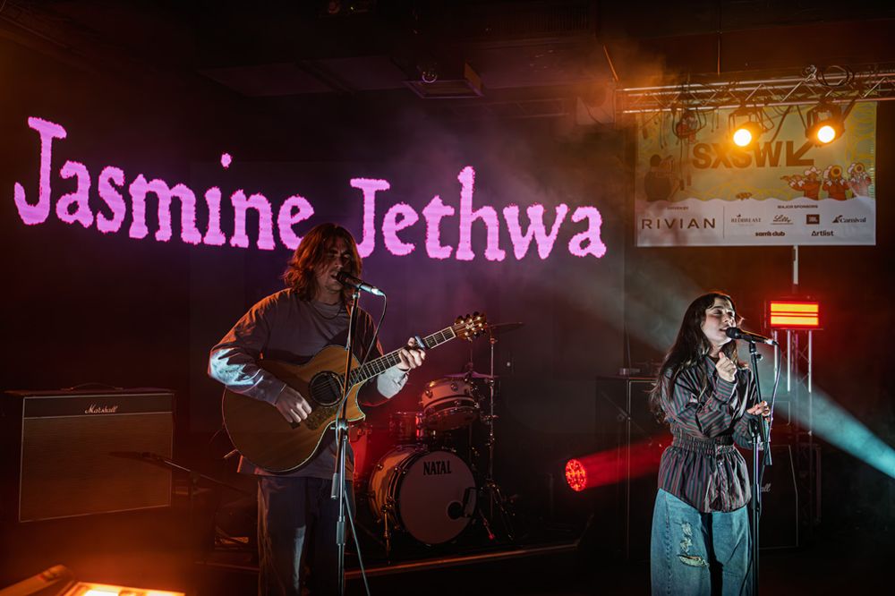 Jasmine Jethwa performing live on stage with guitarist under purple lights and branded backdrop.