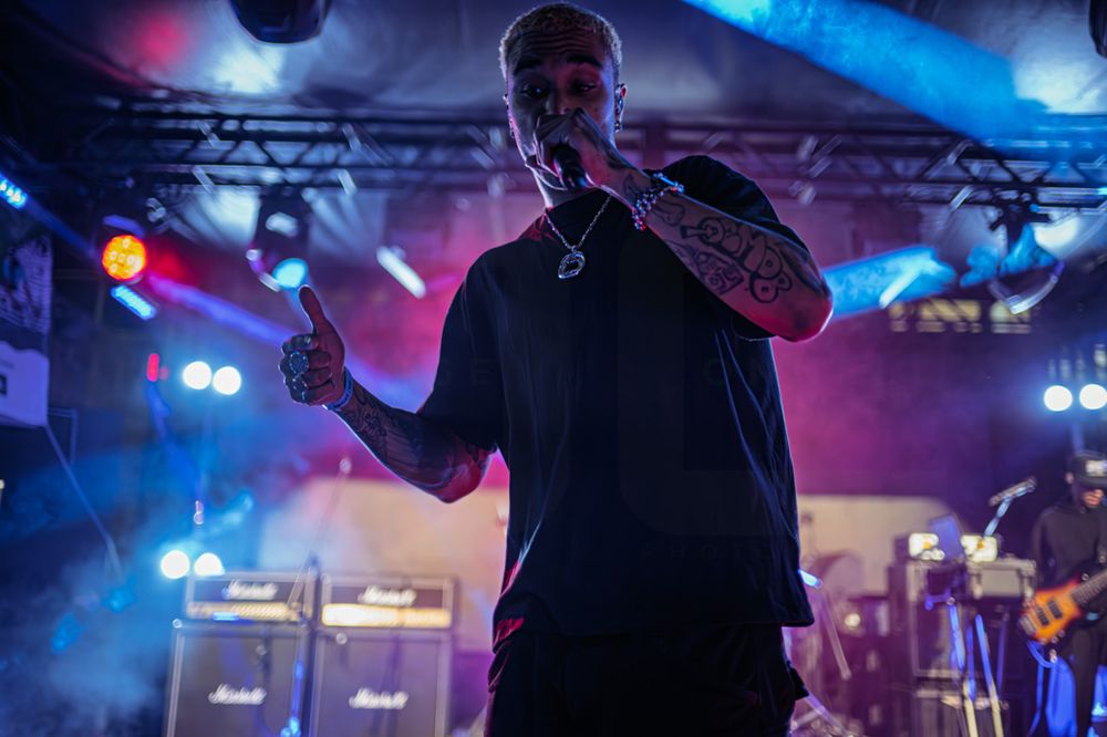 Tattooed rock singer performing on stage under colorful lights with Marshall amplifiers in the background.