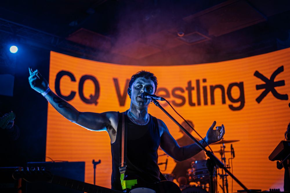 Energetic performer singing on stage at CQ Wrestling event, illuminated by vibrant orange and blue lights.