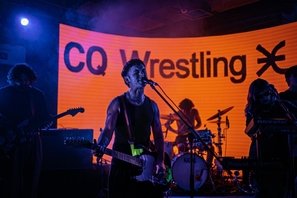 Band performing live on stage in front of a CQ Wrestling sign with dramatic blue and orange lighting.
