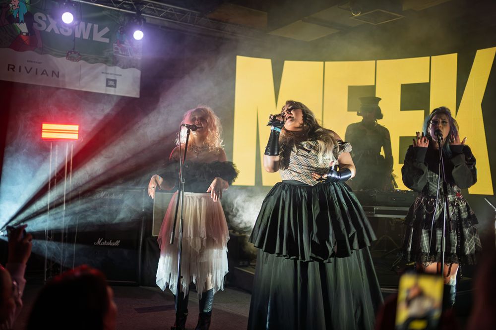 Three women perform on stage at SXSW music festival under dramatic lighting with a large MEEK backdrop.