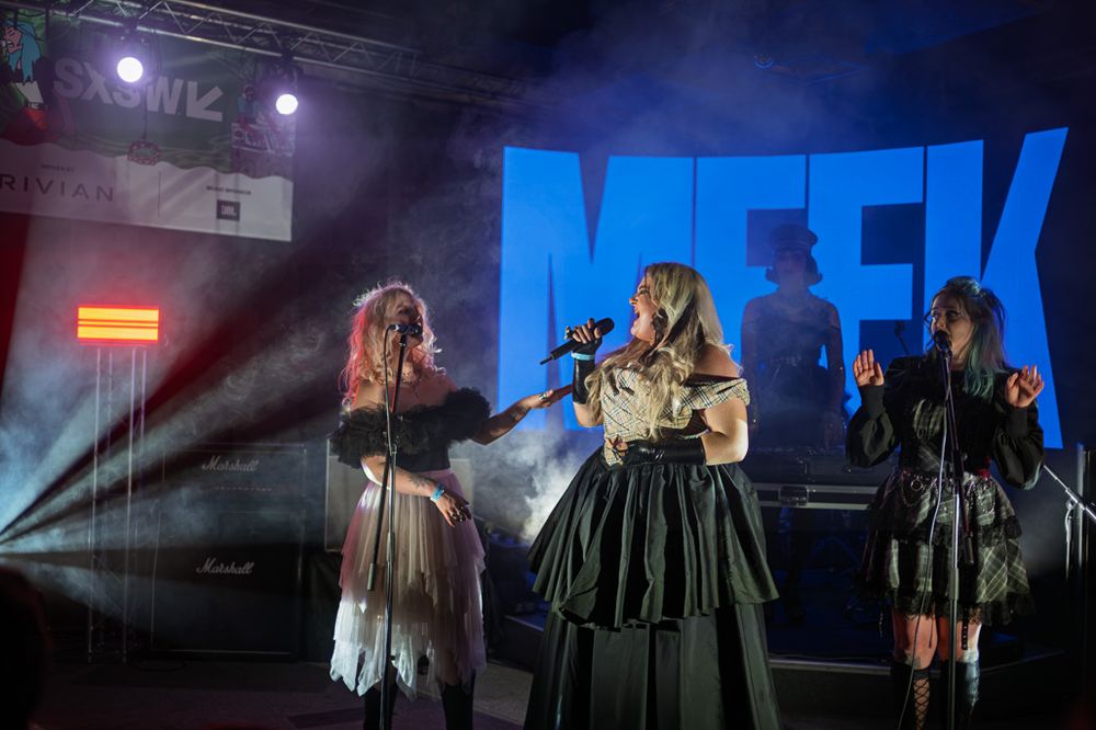 Three female performers on stage at SXSW with dramatic lighting and a large MEEK sign backdrop.