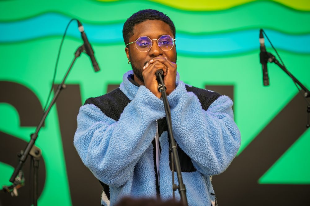 A performer in a blue fleece jacket and purple glasses sings into a microphone on an outdoor stage.