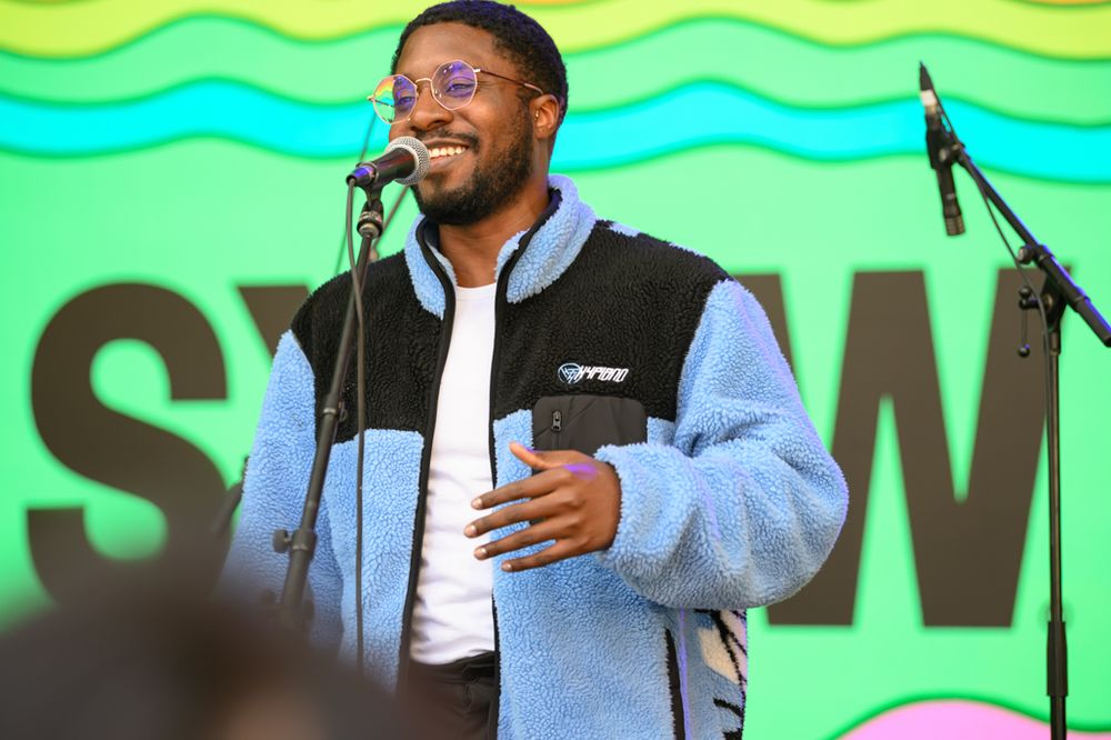 Young man in blue sherpa vest speaking at outdoor music festival microphone on green stage background.