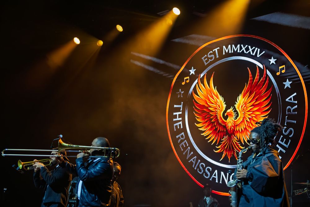 Brass musicians play trombone on a dimly lit stage beneath golden spotlights and a glowing phoenix emblem reading The Renaissance Orchestra.