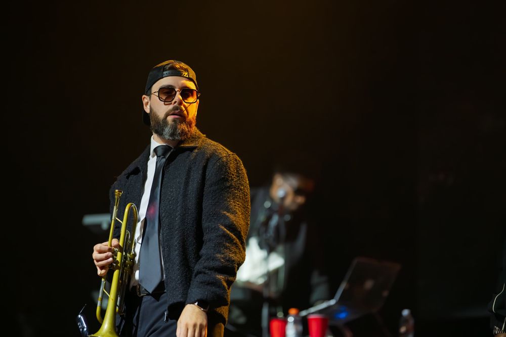 Bearded trumpeter in sunglasses, a cap and a gray jacket holds his trumpet on a dark stage during a live concert performance.