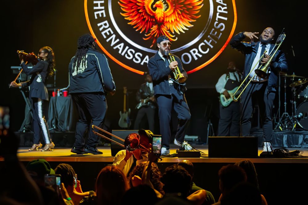 Horn players and guitarists perform on stage beneath a phoenix logo banner while an audience watches and films the live concert.