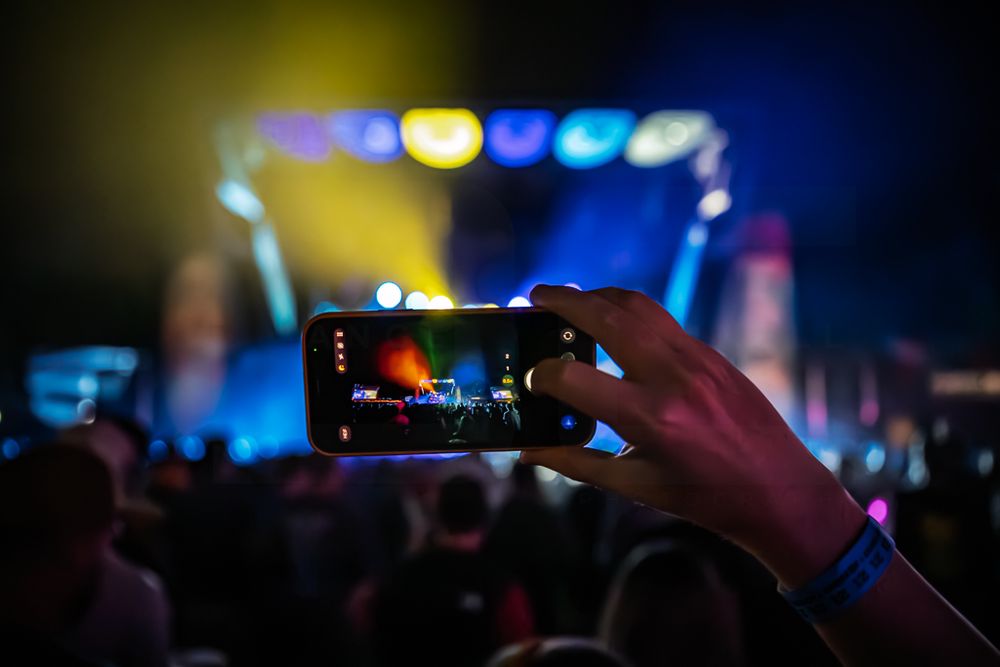 Hand holding a smartphone to record a live concert as colorful stage lights glow above a silhouetted crowd at night.