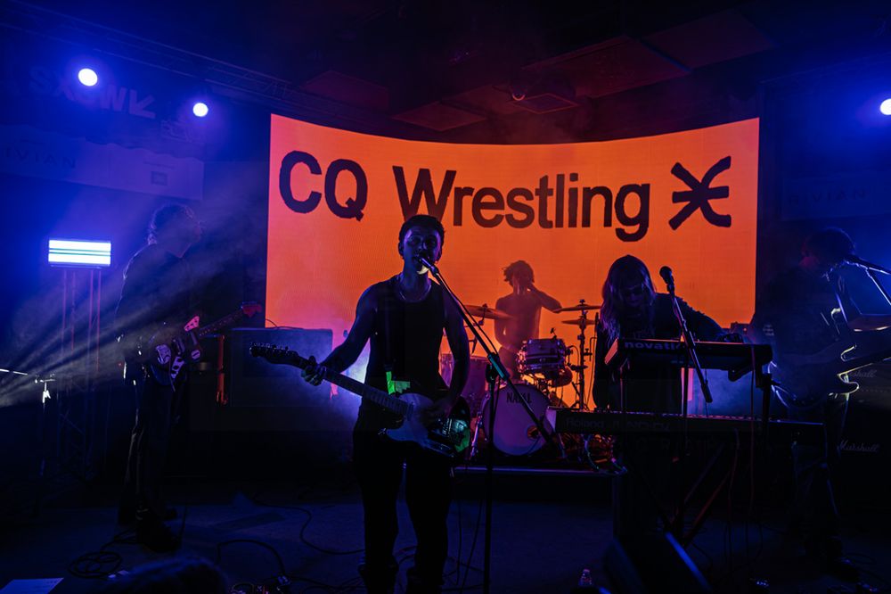 Band performing live on stage in front of an orange CQ Wrestling backdrop with dramatic blue stage lighting.