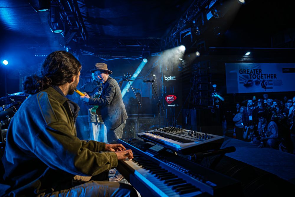 Musician playing keyboard on a blue-lit stage while a vocalist performs for a crowd at a live music venue.