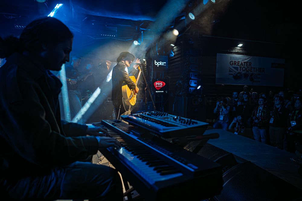Musician plays keyboards on a dimly lit blue-toned stage while a guitarist performs for a crowd at a live concert.