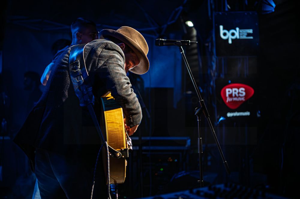 Musician in a wide-brim hat playing acoustic guitar on a dimly lit stage at a PRS for Music event.