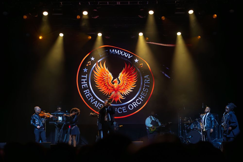 Orchestra musicians perform on a dark stage beneath a glowing phoenix emblem logo as spotlights beam through the haze.