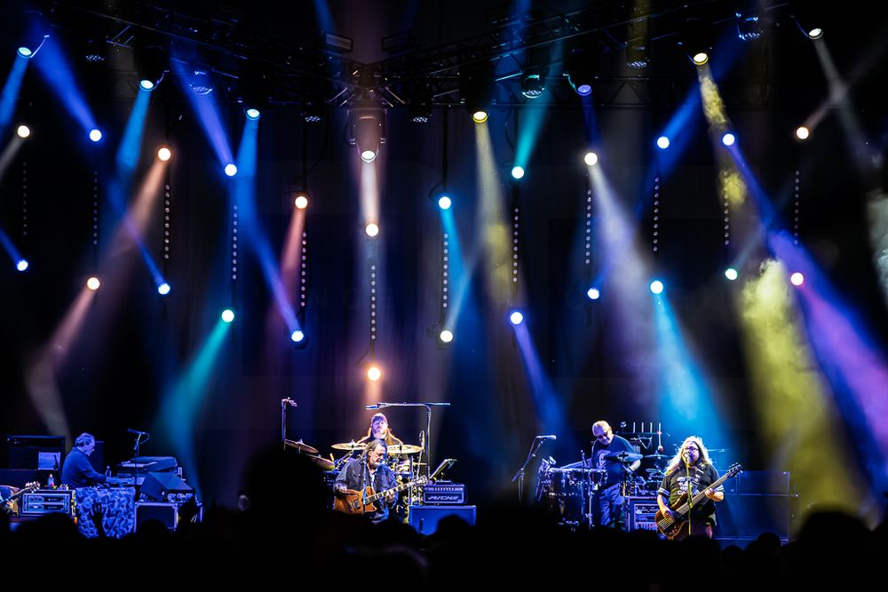 Band performing on a concert stage beneath beams of blue, purple, and yellow lights while a silhouetted crowd watches.