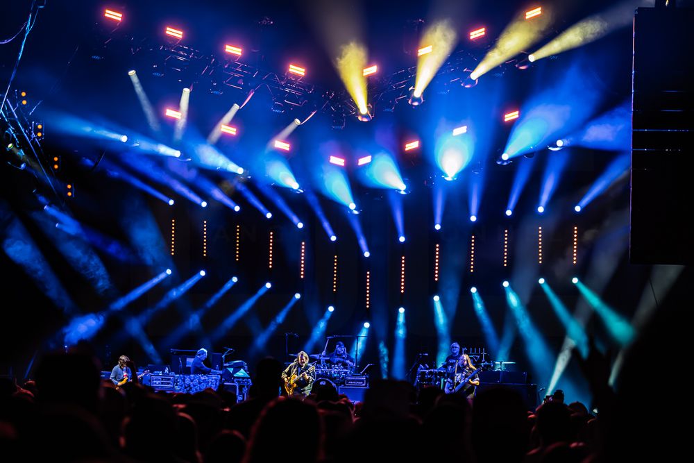 Band performing on a dark concert stage as vibrant blue and amber spotlight beams fill the venue above a silhouetted crowd.