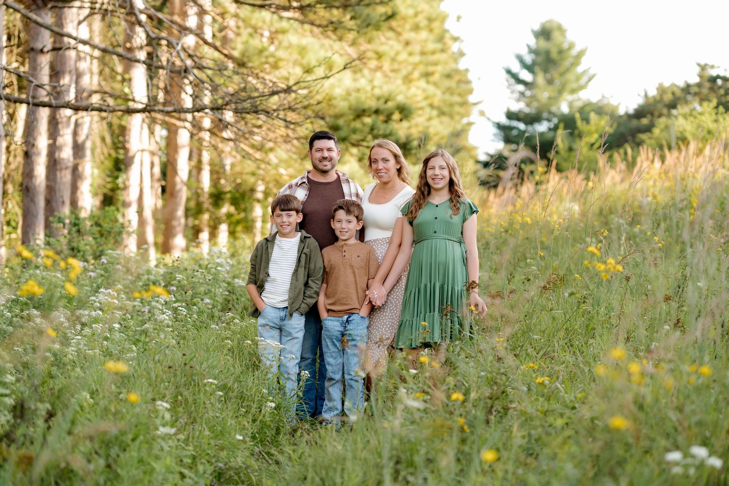 A happy family of five poses together in a sunny wildflower meadow surrounded by tall green trees.